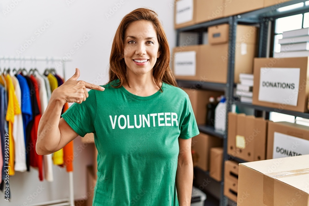 Middle age hispanic woman wearing volunteer t shirt at donations stand pointing finger to one self smiling happy and proud Middle age hispanic woman wearing volunteer t shirt at donations stand pointing finger to one self smiling happy and proud