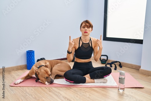 Young beautiful woman sitting on yoga mat pointing up looking sad and upset, indicating direction with fingers, unhappy and depressed.