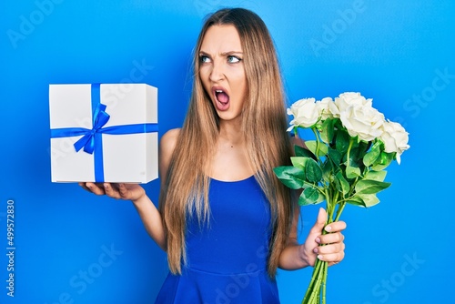 Young blonde girl holding anniversary present and bouquet of flowers angry and mad screaming frustrated and furious, shouting with anger. rage and aggressive concept.