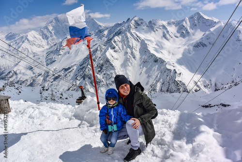 A little boy and a man on the background of snow-capped mountains and the flag of Russia. Elbrus April 25, 2019.