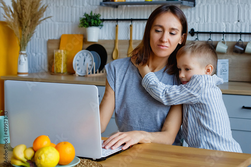 Working mother concept. Young woman working on laptop with her child from home. Stay at home mom working remotely on laptop