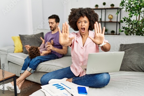 Mother of interracial family working using computer laptop at home doing stop gesture with hands palms, angry and frustration expression
