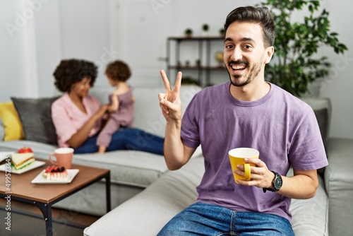 Hispanic father of interracial family drinking a cup coffee smiling with happy face winking at the camera doing victory sign with fingers. number two.