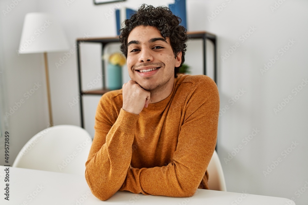 Young hispanic man smiling happy sitting on the table at home. Young hispanic man smiling happy sitting on the table at home.