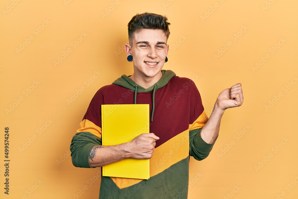 Young caucasian boy with ears dilation holding book screaming proud, celebrating victory and success very excited with raised arm Young caucasian boy with ears dilation holding book screaming proud, celebrating victory and success very excited with raised arm