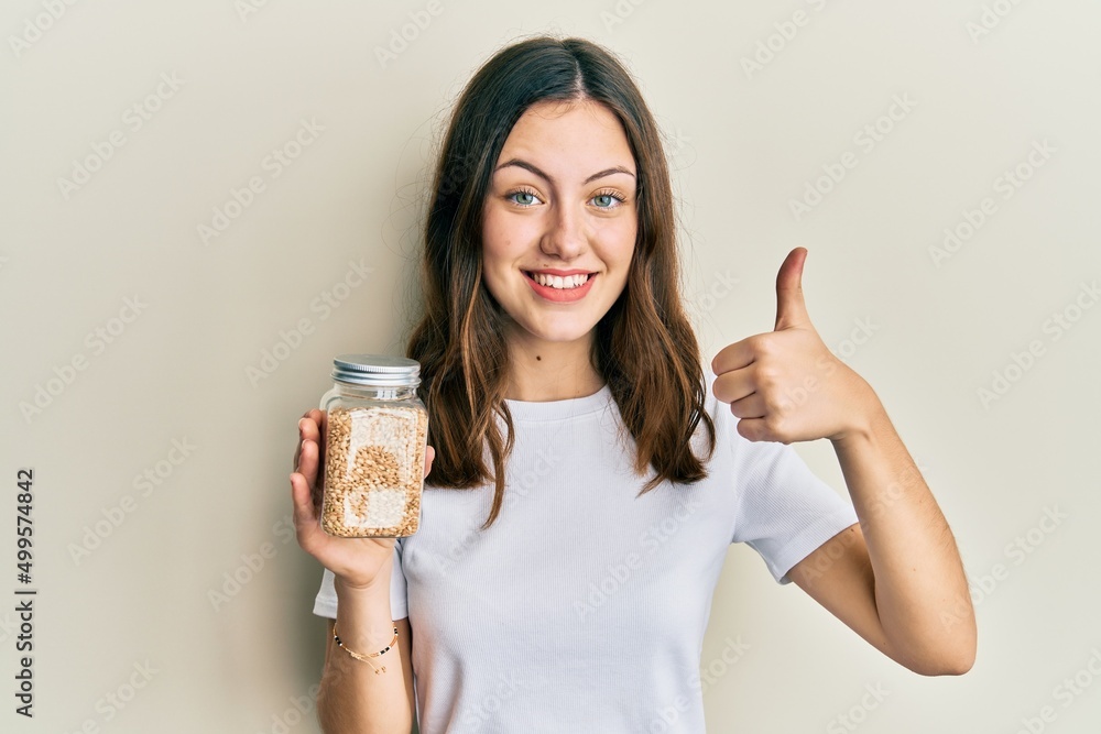 Young brunette woman holding jar with rice smiling happy and positive, thumb up doing excellent and approval sign Young brunette woman holding jar with rice smiling happy and positive, thumb up doing excellent and approval sign