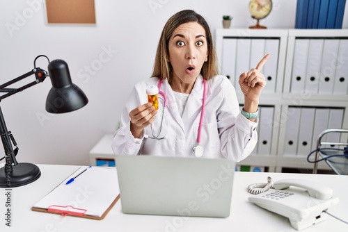 Young hispanic woman wearing doctor uniform holding pills at the clinic surprised pointing with finger to the side  open mouth amazed expression.