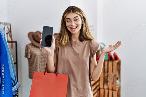 Young blonde woman holding shopping bags showing smartphone screen celebrating achievement with happy smile and winner expression with raised hand