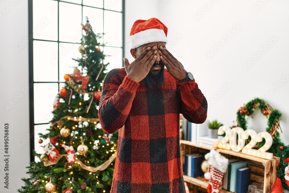 African american man wearing santa claus hat standing by christmas tree rubbing eyes for fatigue and headache, sleepy and tired expression. vision problem