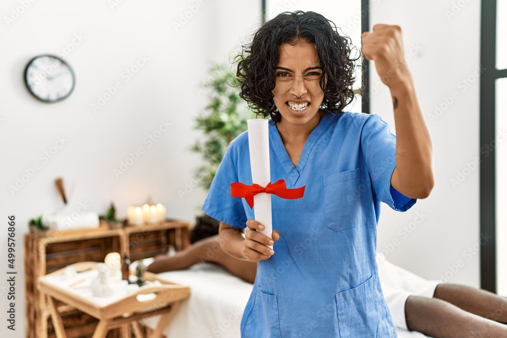 Young therapist woman holding diploma at spa wellness center annoyed and frustrated shouting with anger, yelling crazy with anger and hand raised