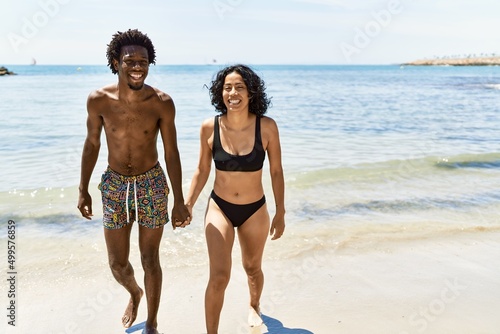 Young interracial tourist couple wearing swimwear walking with hands together at the beach.