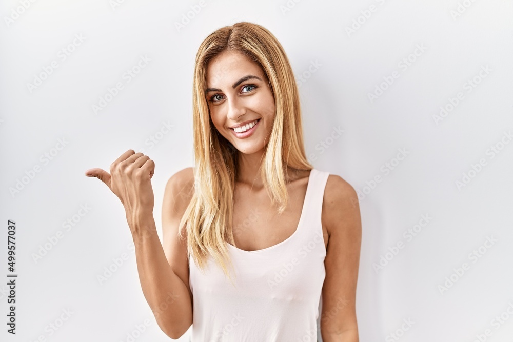 Blonde beautiful young woman standing over white isolated background smiling with happy face looking and pointing to the side with thumb up. Blonde beautiful young woman standing over white isolated background smiling with happy face looking and pointing to the side with thumb up.