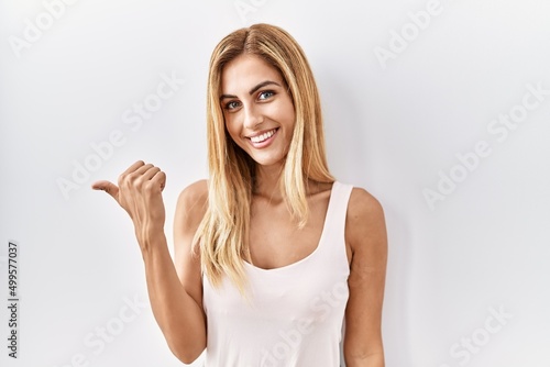 Blonde beautiful young woman standing over white isolated background smiling with happy face looking and pointing to the side with thumb up.
