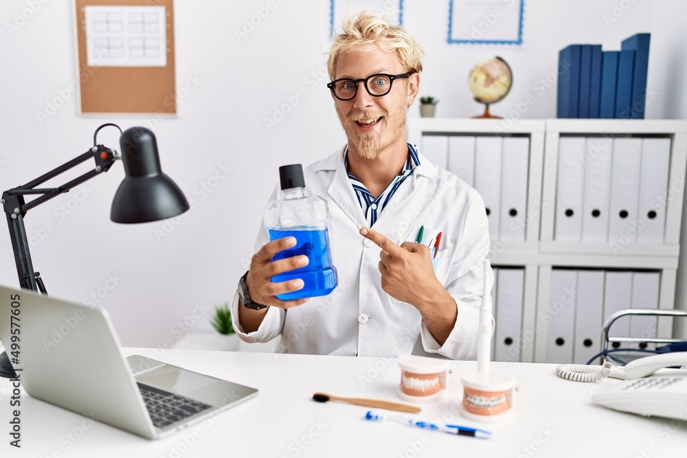 Young blond dentist man holding mouthwash at clinic smiling happy pointing with hand and finger Young blond dentist man holding mouthwash at clinic smiling happy pointing with hand and finger