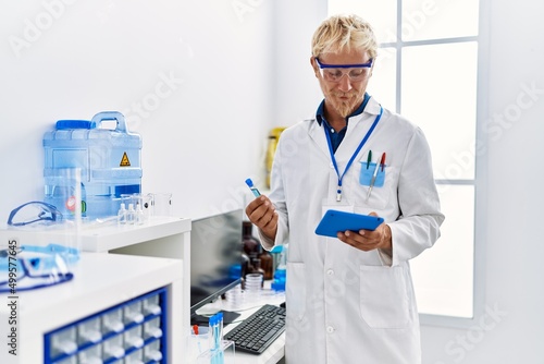 Young caucasian man wearing scientist uniform holding test tube and touchpad at laboratory