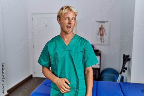 Young blond man wearing physiotherapist uniform standing at clinic winking looking at the camera with sexy expression, cheerful and happy face.
