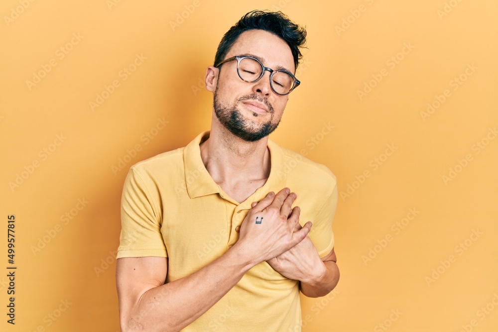 Young hispanic man wearing casual clothes and glasses smiling with hands on chest with closed eyes and grateful gesture on face. health concept. Young hispanic man wearing casual clothes and glasses smiling with hands on chest with closed eyes and grateful gesture on face. health concept.