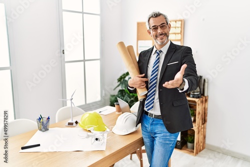 Middle age hispanic business man holding paper blueprints smiling cheerful offering palm hand giving assistance and acceptance.
