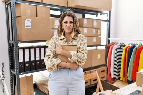 Young hispanic shopkeeper woman holding cardboard box at storeware clueless and confused expression. doubt concept. photo