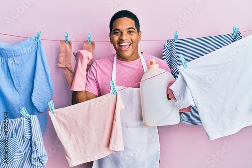 Young handsome hispanic man doing laundry holding detergent bottle pointing thumb up to the side smiling happy with open mouth