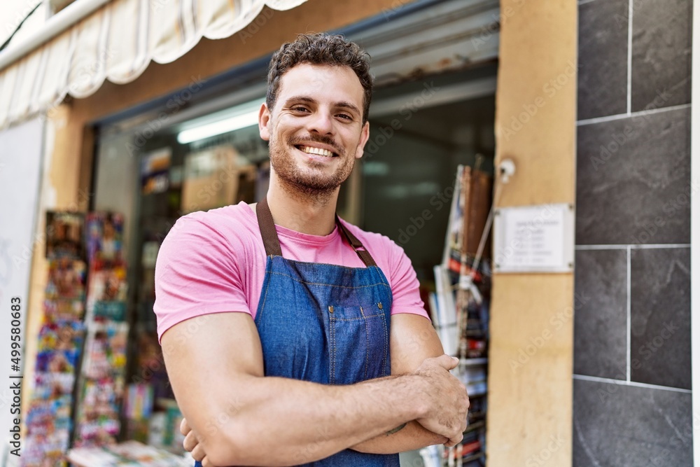 Fototapeta premium Young hispanic worker wearing apron standing with arms crossed gesture at newspaper store.