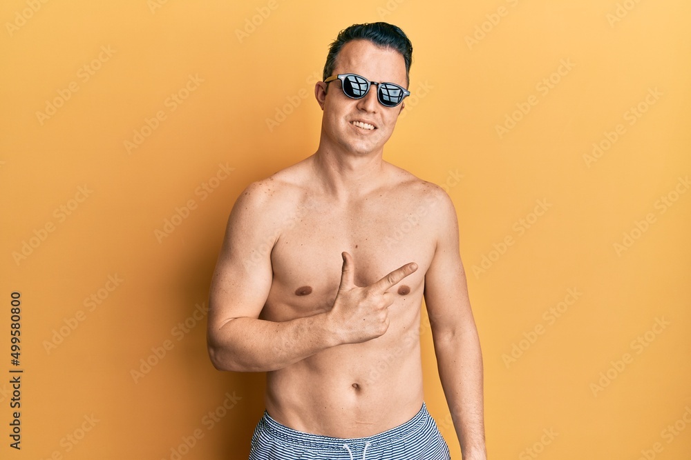 Fototapeta premium Handsome young man wearing swimsuit and sunglasses smiling cheerful pointing with hand and finger up to the side