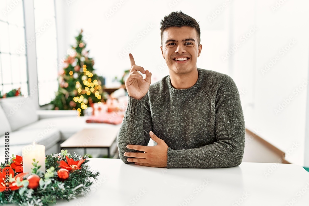 Young hispanic man sitting on the table by christmas tree showing and pointing up with finger number one while smiling confident and happy. Young hispanic man sitting on the table by christmas tree showing and pointing up with finger number one while smiling confident and happy.