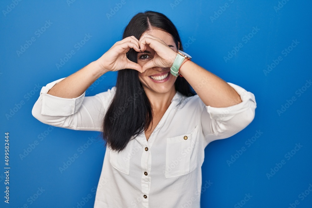 Young hispanic woman standing over blue background doing heart shape with hand and fingers smiling looking through sign Young hispanic woman standing over blue background doing heart shape with hand and fingers smiling looking through sign