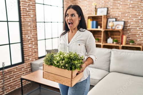 Young hispanic woman holding green plants at the living room angry and mad screaming frustrated and furious  shouting with anger. rage and aggressive concept.