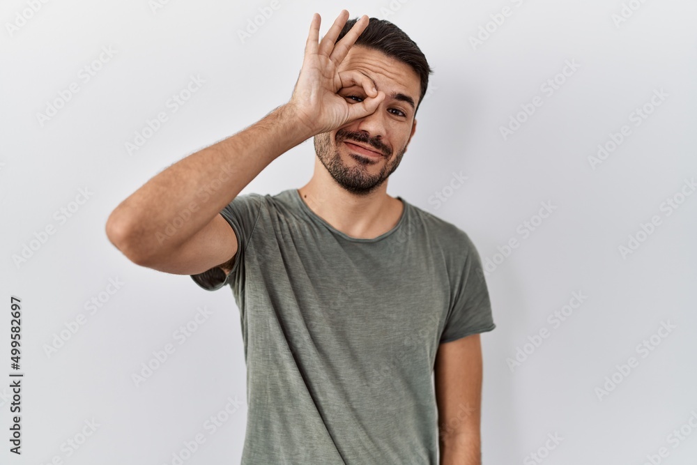 Young hispanic man with beard wearing casual t shirt over white background doing ok gesture with hand smiling, eye looking through fingers with happy face. Young hispanic man with beard wearing casual t shirt over white background doing ok gesture with hand smiling, eye looking through fingers with happy face.