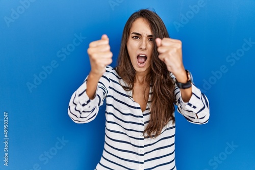 Young hispanic woman standing over blue isolated background angry and mad raising fists frustrated and furious while shouting with anger. rage and aggressive concept.