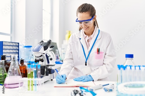 Young hispanic woman wearing scientist uniform writing on notebook using microscope at laboratory
