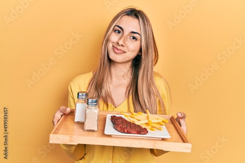 Beautiful hispanic woman holding tray with lunch relaxed with serious expression on face. simple and natural looking at the camera.