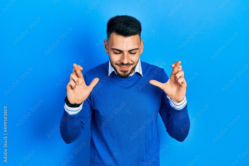 Young hispanic man with beard wearing casual blue sweater gesturing finger crossed smiling with hope and eyes closed. luck and superstitious concept. Young hispanic man with beard wearing casual blue sweater gesturing finger crossed smiling with hope and eyes closed. luck and superstitious concept.