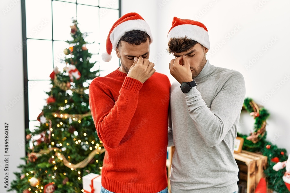 Young gay couple standing by christmas tree wearing hat tired rubbing nose and eyes feeling fatigue and headache. stress and frustration concept. Young gay couple standing by christmas tree wearing hat tired rubbing nose and eyes feeling fatigue and headache. stress and frustration concept.