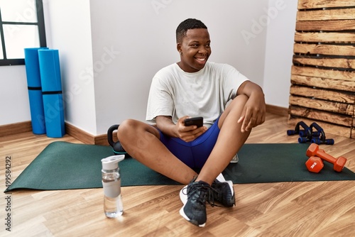 Young african man sitting on training mat at the gym using smartphone looking away to side with smile on face, natural expression. laughing confident.