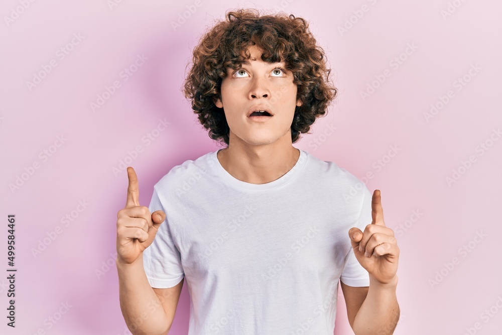 Handsome young man wearing casual white t shirt amazed and surprised looking up and pointing with fingers and raised arms. Handsome young man wearing casual white t shirt amazed and surprised looking up and pointing with fingers and raised arms.