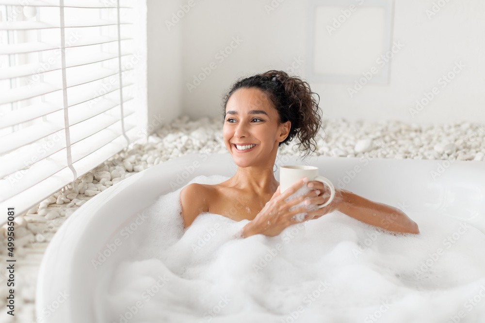 Naklejka premium Carefree young woman sitting in foamy bath, drinking coffee or tea near window at home, free space