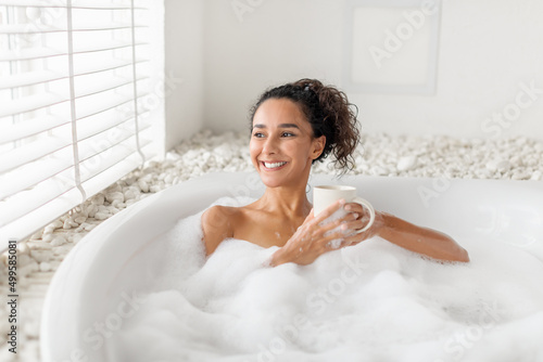 Carefree young woman sitting in foamy bath, drinking coffee or tea near window at home, free space