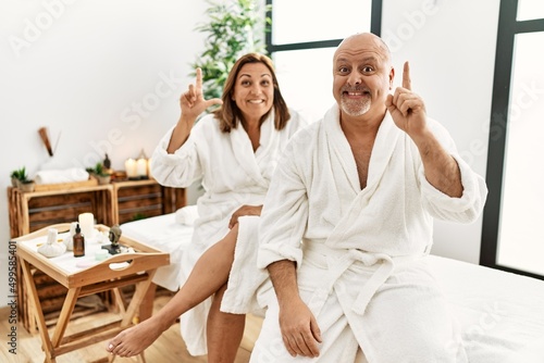 Middle age hispanic couple wearing bathrobe at wellness spa pointing finger up with successful idea. exited and happy. number one.