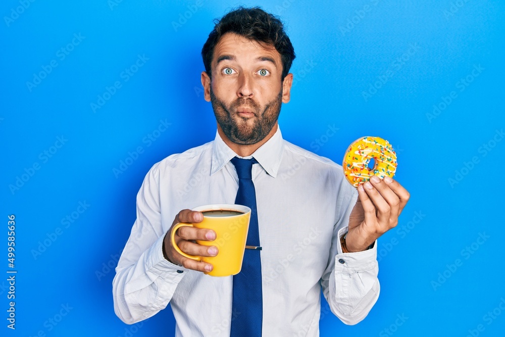 Handsome business man with beard eating doughnut and drinking coffee making fish face with mouth and squinting eyes, crazy and comical. Handsome business man with beard eating doughnut and drinking coffee making fish face with mouth and squinting eyes, crazy and comical.