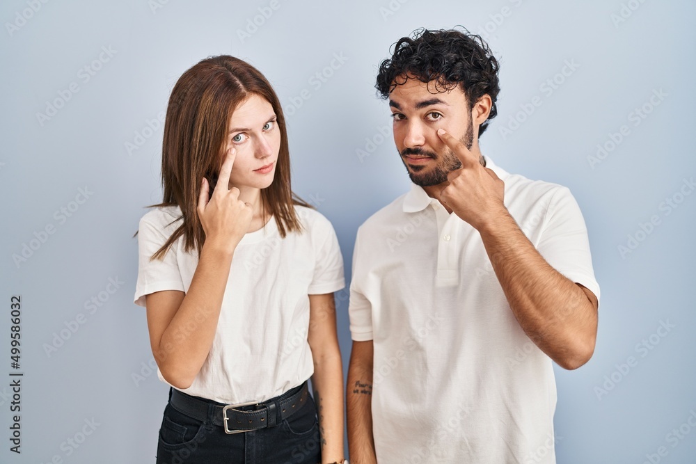 Young couple wearing casual clothes standing together pointing to the eye watching you gesture, suspicious expression Young couple wearing casual clothes standing together pointing to the eye watching you gesture, suspicious expression