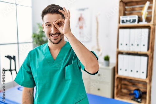 Young physiotherapist man working at pain recovery clinic doing ok gesture with hand smiling, eye looking through fingers with happy face.