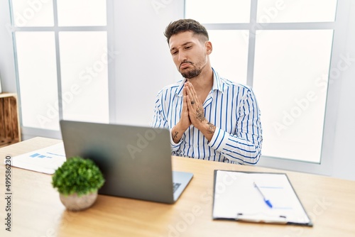 Young handsome man with beard working at the office using computer laptop begging and praying with hands together with hope expression on face very emotional and worried. begging.