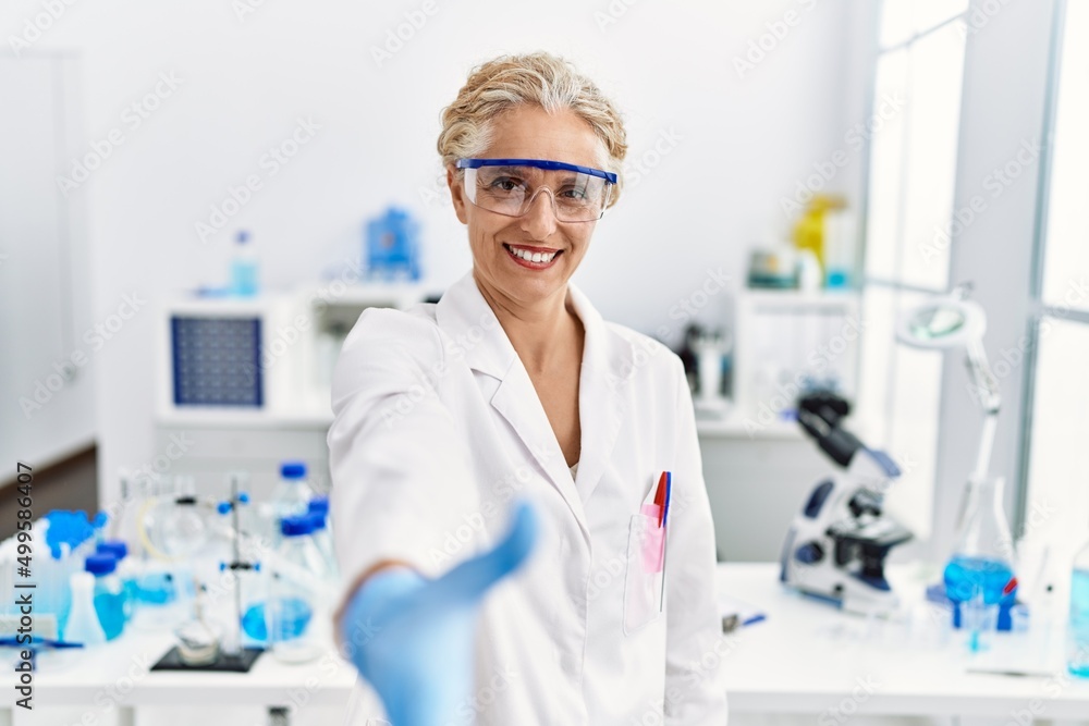 Middle age blonde woman working at scientist laboratory smiling friendly offering handshake as greeting and welcoming. successful business.