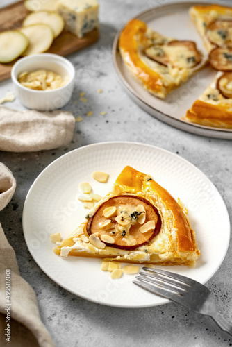 A slice of puff pastry pizza with pear, almonds, gorgonzola ricotta or blue cheese in a white plate on the kitchen table. Delicious layered pie with pear, dorblu, nuts on a light culinary background