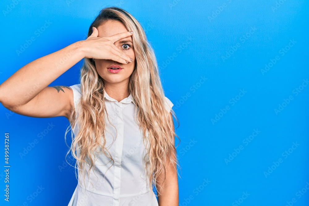 Beautiful young blonde woman wearing casual white shirt peeking in shock covering face and eyes with hand, looking through fingers with embarrassed expression. Beautiful young blonde woman wearing casual white shirt peeking in shock covering face and eyes with hand, looking through fingers with embarrassed expression.