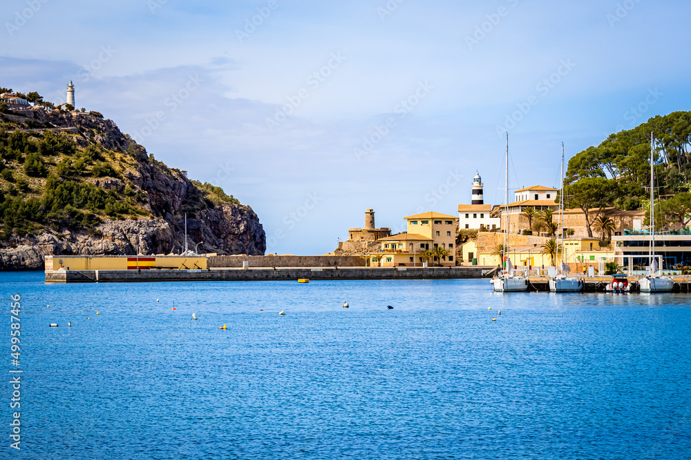 Photography of the tranquil harbor basin of port de soller with three sailboats and one motorboat moored at the pier. Lighthouse Far de sa creu and beacon far del cap gros in the background.