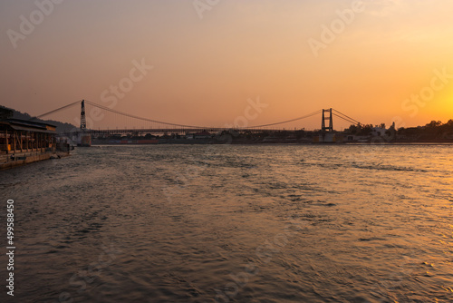 iron suspension bridge with sunset orange sky over flowing river horizon at evening from flat angle