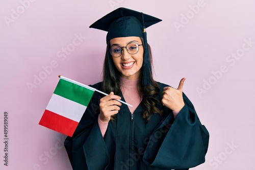 Young hispanic woman wearing graduation uniform holding italy flag smiling happy and positive, thumb up doing excellent and approval sign